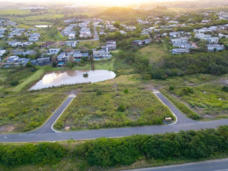 Aerial view of a vacant residential plot featuring paved road access and proximity to a scenic estate pond and surrounding neighborhood.