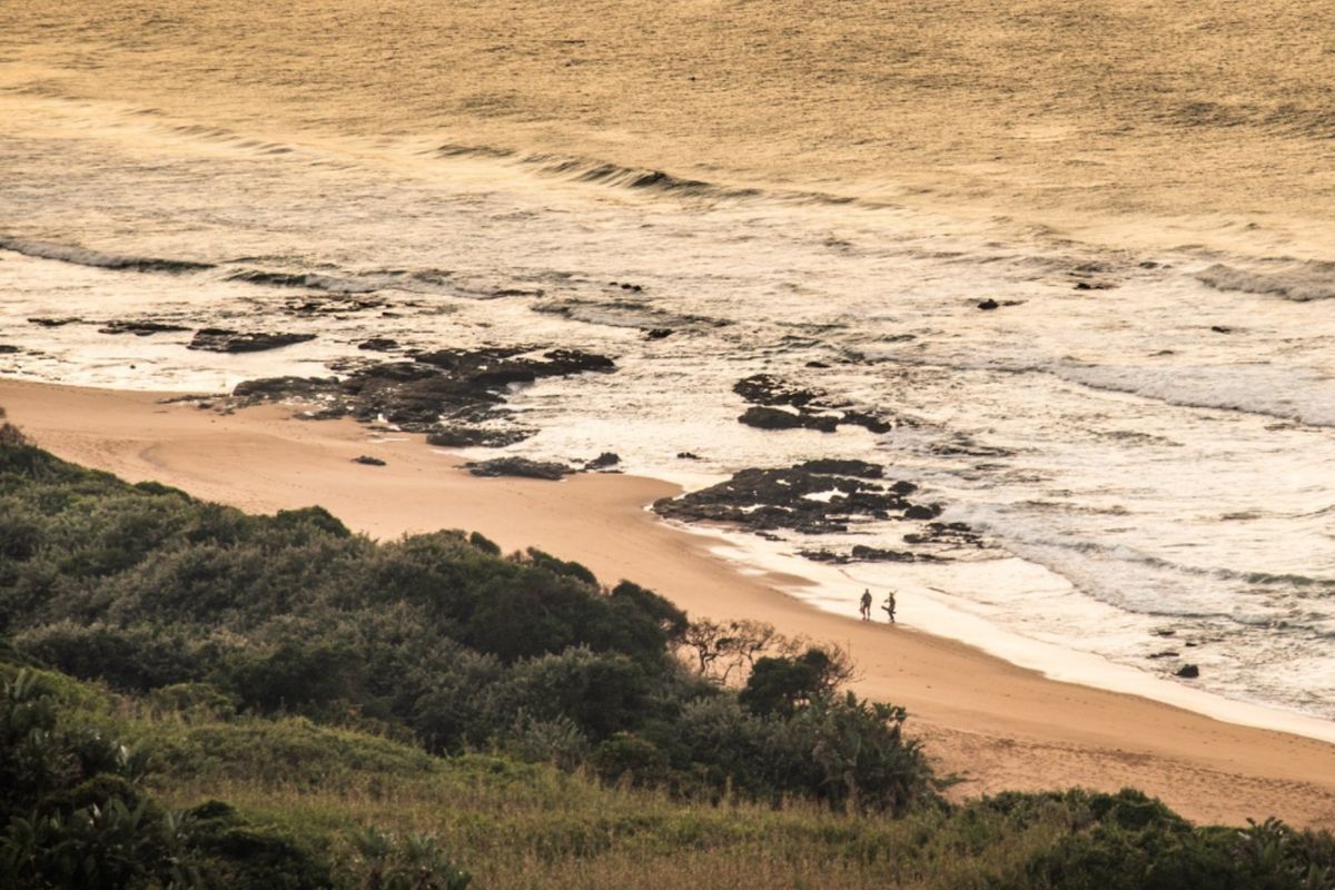 A scenic elevated view of the coastline, featuring sandy beach, rocky outcrops, and ocean waves at sunset.