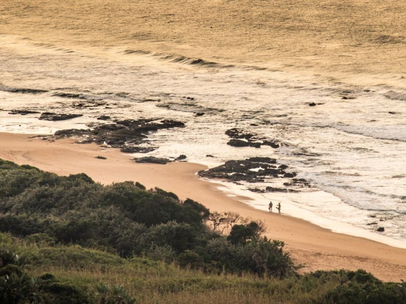 A scenic elevated view of the coastline, featuring sandy beach, rocky outcrops, and ocean waves at sunset.