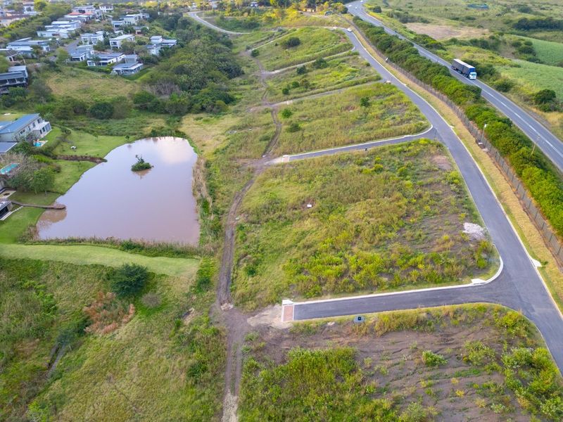 Aerial view of a vacant residential plot featuring natural landscape, a pond, and established paved road access within a developing estate.