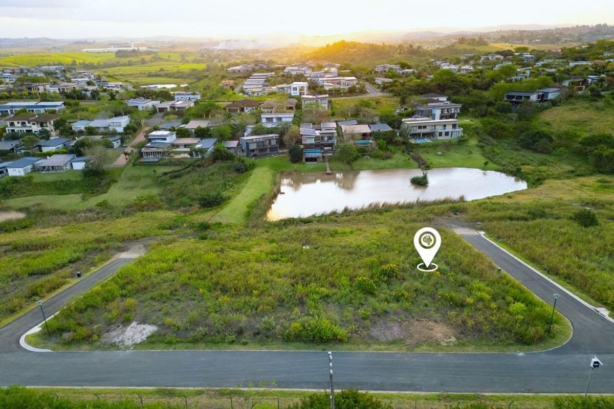 Aerial view of a vacant residential plot in a developed estate, featuring road access and proximity to a scenic pond and surrounding neighborhood.