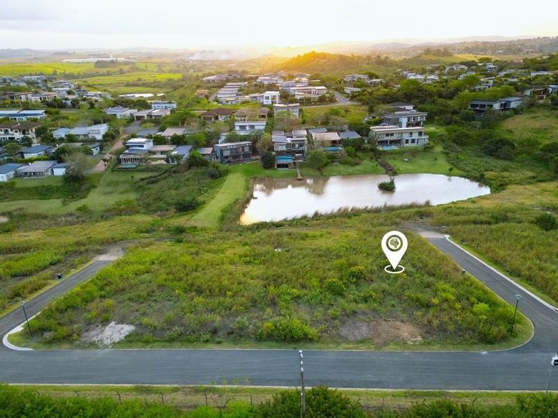 Aerial view of a vacant residential plot in a developed estate, featuring road access and proximity to a scenic pond and surrounding neighborhood.