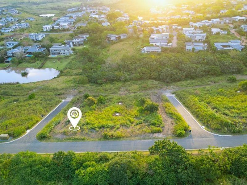 An aerial view of a vacant residential plot situated within a developed estate, featuring road access and surrounding greenery.