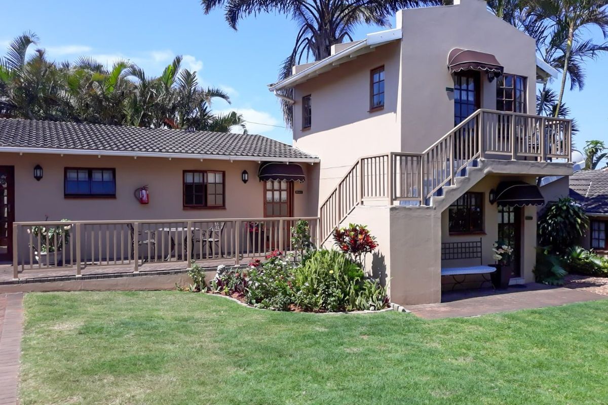 A well-maintained guest house exterior featuring a timber deck, external staircase leading to a balcony, and landscaped garden area.