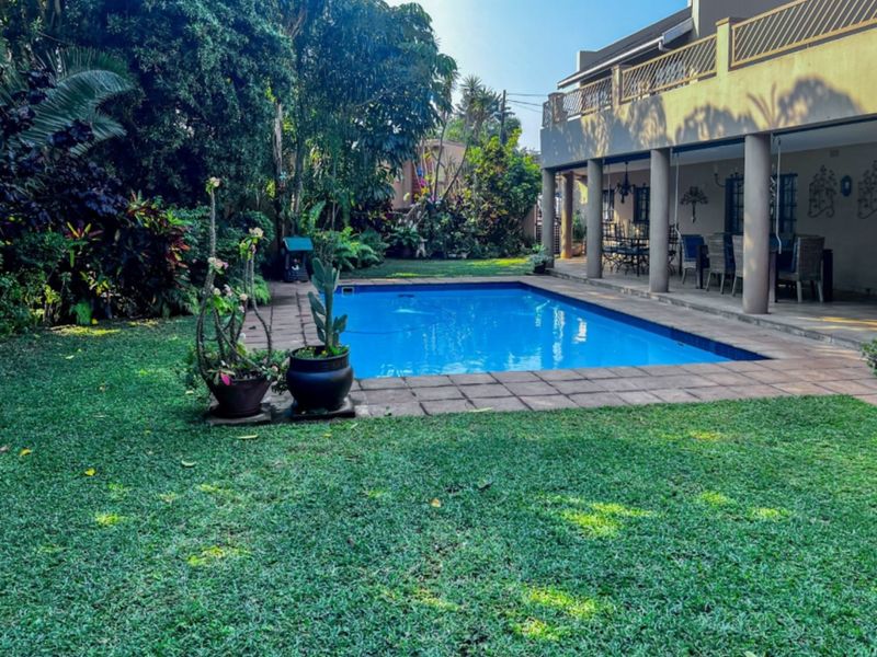 A well-maintained swimming pool area featuring a paved deck, lush garden surroundings, and a spacious covered patio attached to the main building.