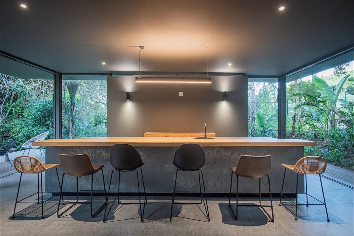 A modern, minimalist kitchen island featuring a stone base, wooden countertop, and integrated sink, set within a space surrounded by floor-to-ceiling glass walls.