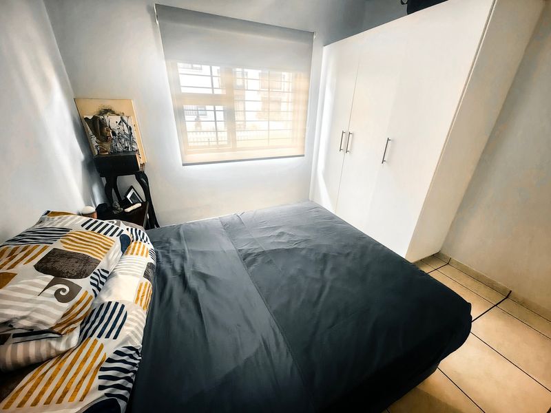 A compact bedroom featuring tiled flooring, a built-in wardrobe, and a window fitted with a roller blind and security bars.