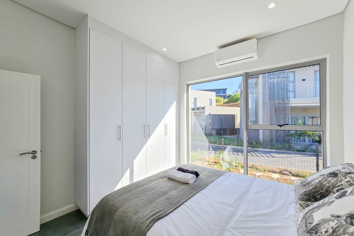 A modern, well-lit bedroom featuring built-in cupboards, a split-system air conditioner, and large windows with views of the surrounding neighbourhood.