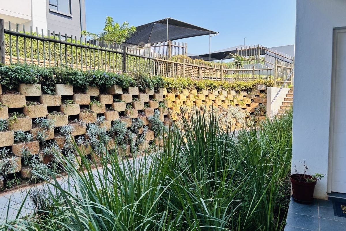 A well-maintained rear exterior area featuring a tiered retaining wall with integrated planting, timber fencing, and a paved walkway leading to a shaded outdoor space.