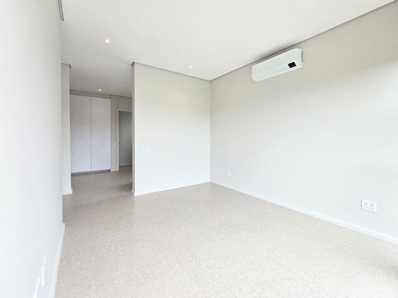 A bright, modern, and empty bedroom featuring neutral tiled flooring, recessed lighting, built-in storage, and a wall-mounted air conditioning unit.