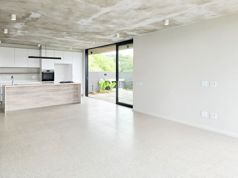 Modern open-plan living area featuring an exposed concrete ceiling, sleek kitchen with island, and large sliding doors opening to an outdoor patio.