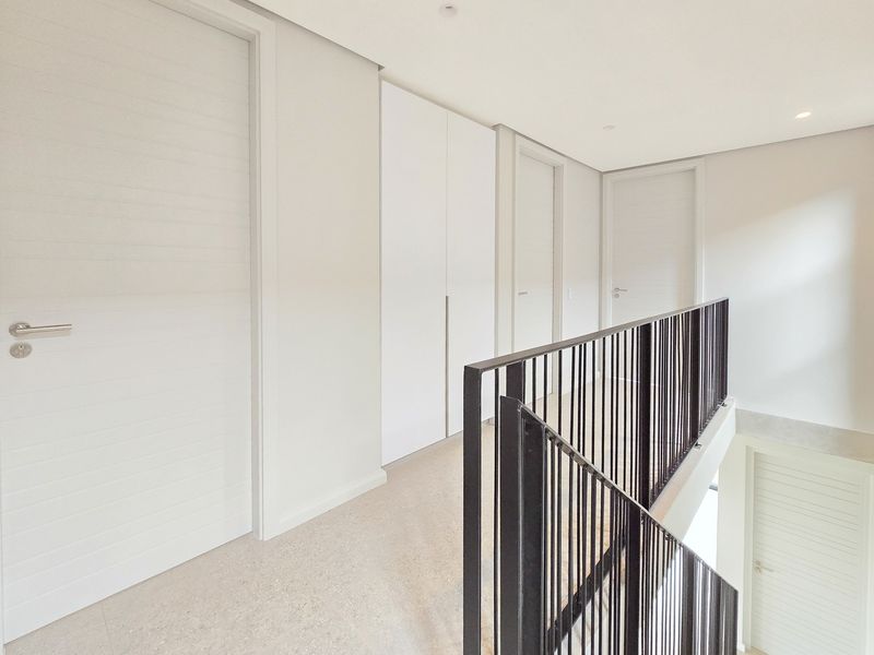 A bright and modern upper-level hallway featuring clean white finishes, built-in storage cupboards, and a sleek black metal balustrade overlooking the lower level.