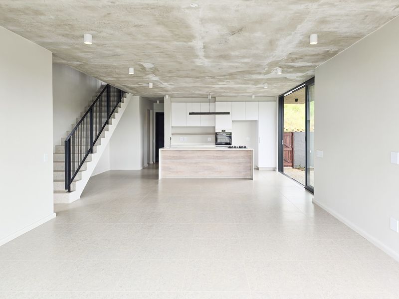 Spacious open-plan living area featuring an exposed concrete ceiling, modern kitchen with island, and a staircase, leading out to a patio through sliding doors.
