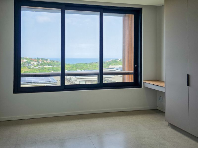 A bright, modern bedroom featuring built-in cupboards, a fitted desk, and large sliding windows offering scenic coastal views.