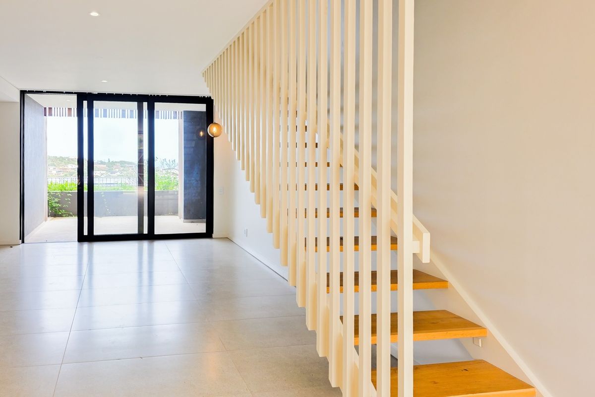 Modern open-plan living area featuring a striking timber staircase with vertical slat balustrade, tiled flooring, and large sliding glass doors leading to a patio.