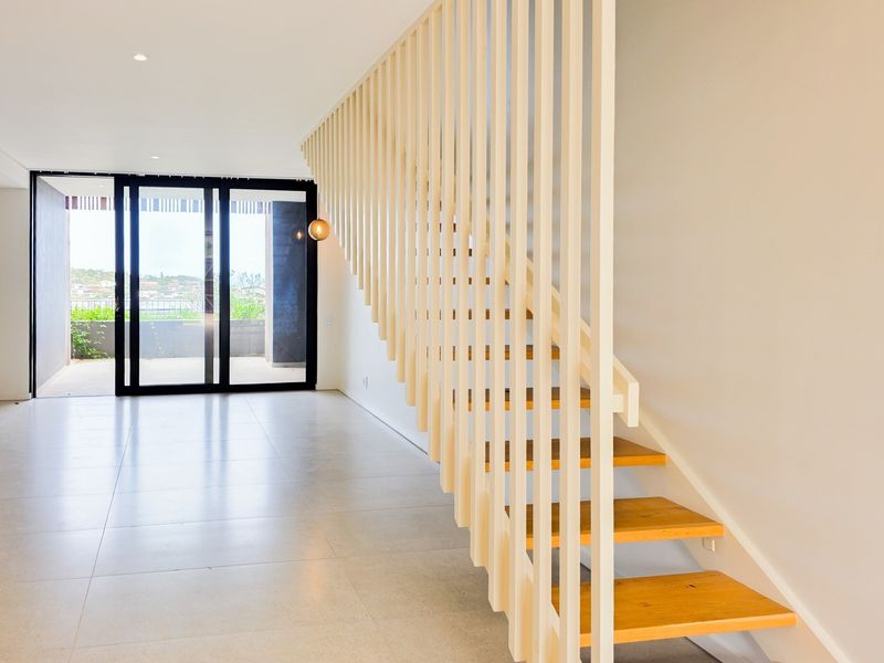 Modern open-plan living area featuring a striking timber staircase with vertical slat balustrade, tiled flooring, and large sliding glass doors leading to a patio.