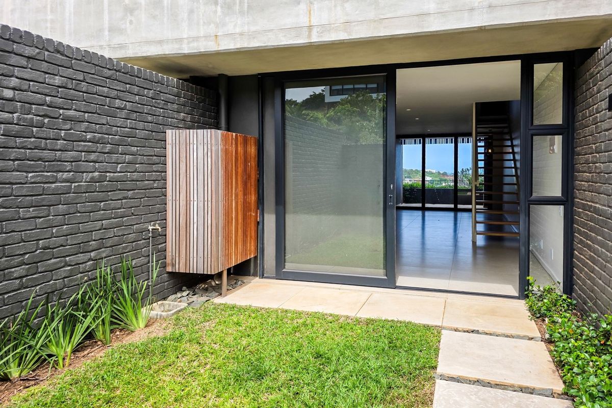 A modern rear exterior view of a townhouse featuring a private garden, paved walkway, and large sliding glass doors leading into the open-plan living area.