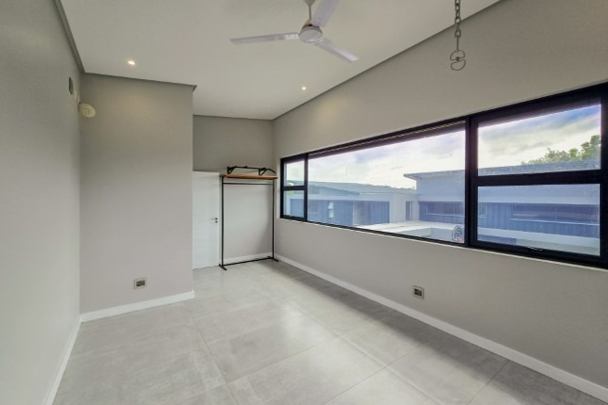 A bright, modern, and empty bedroom featuring large horizontal windows, neutral tiled flooring, recessed lighting, and a ceiling fan.