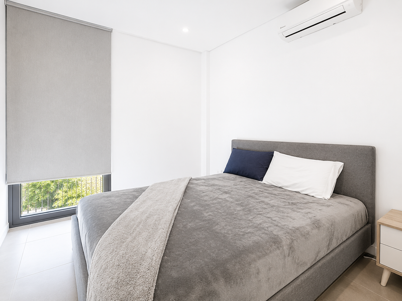 Modern bedroom featuring neutral tiled flooring, a wall-mounted split-system air conditioner, and a large window with a fitted roller blind.