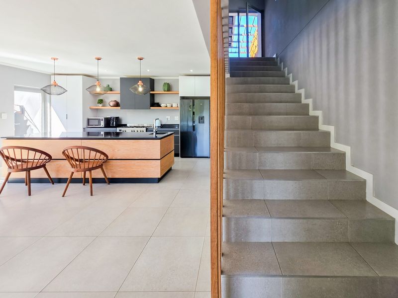 Modern open-plan kitchen featuring a central island, sleek cabinetry, and tiled flooring, adjacent to a contemporary staircase with a wooden handrail.