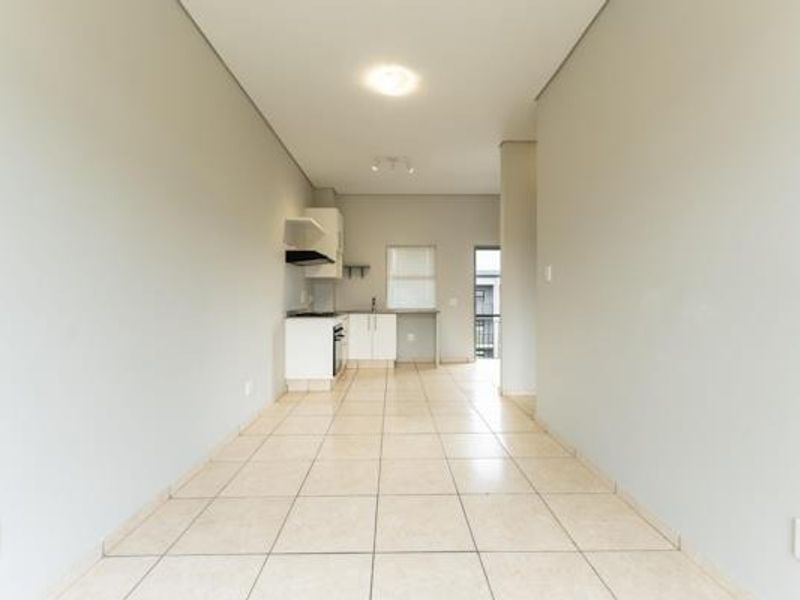 An open-plan living and kitchen area featuring neutral tiled flooring, built-in cabinetry, and integrated kitchen appliances in a clean, vacant space.