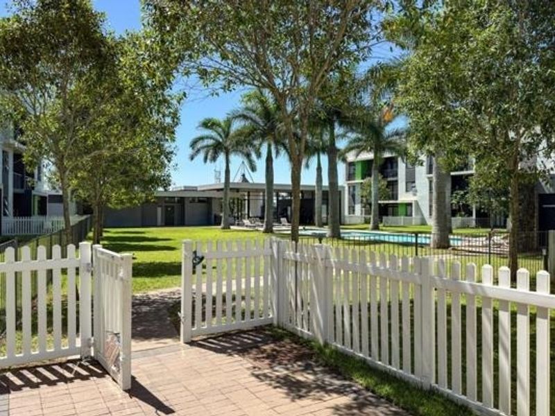 A view of the apartment complex communal pool area, featuring landscaped gardens, mature palm trees, and a white picket fence enclosure.