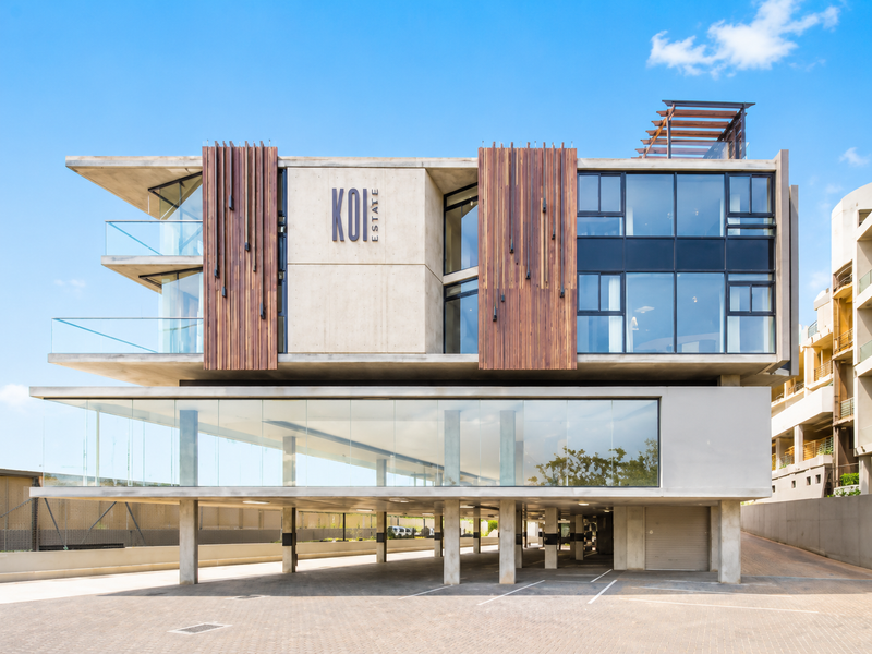 Modern multi-story apartment building featuring contemporary concrete and timber architecture, glass balconies, and a spacious ground-level covered parking area.