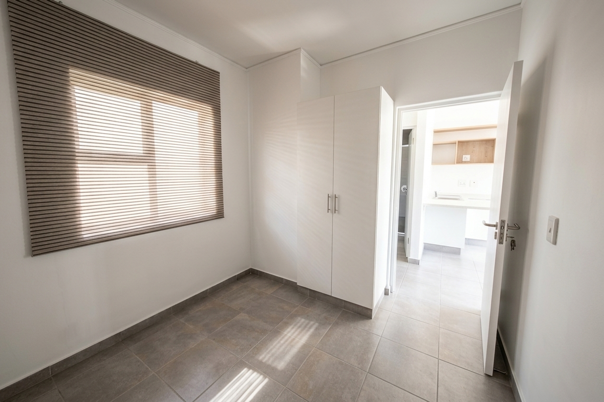 A bright, empty bedroom featuring neutral tiled flooring, a built-in wardrobe, and venetian blinds, with an open doorway leading to an adjacent kitchen area.