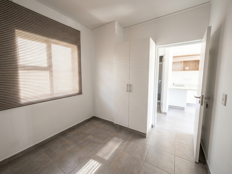 A bright, empty bedroom featuring neutral tiled flooring, a built-in wardrobe, and venetian blinds, with an open doorway leading to an adjacent kitchen area.
