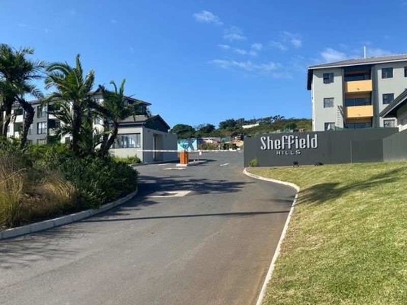 The entrance to Sheffield Hills apartment complex featuring a secure boom gate, paved driveway, and well-maintained landscaped gardens.
