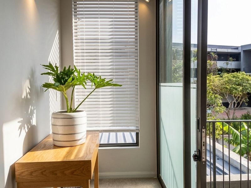 A bright, modern balcony area featuring a sliding glass door, metal balustrade, and venetian blinds, offering a pleasant view of the surrounding complex.