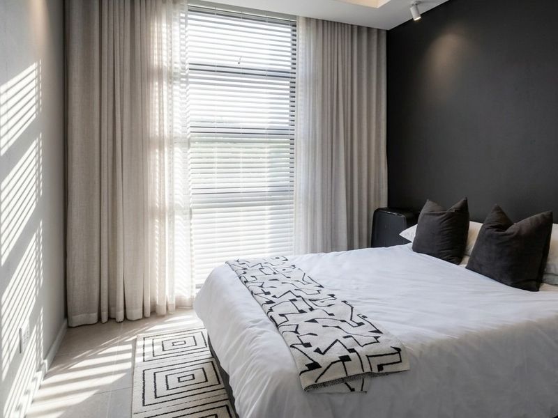 A modern, well-lit bedroom featuring neutral tiled flooring, a large window with integrated blinds, and contemporary dark accent wall detailing.