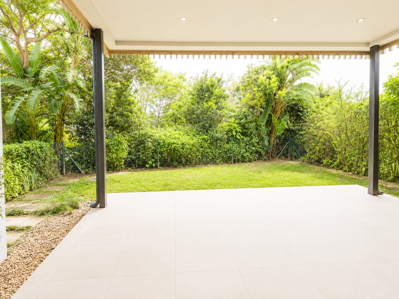 A spacious, modern covered patio featuring light-colored floor tiles, recessed lighting, and direct access to a lush, private garden area.