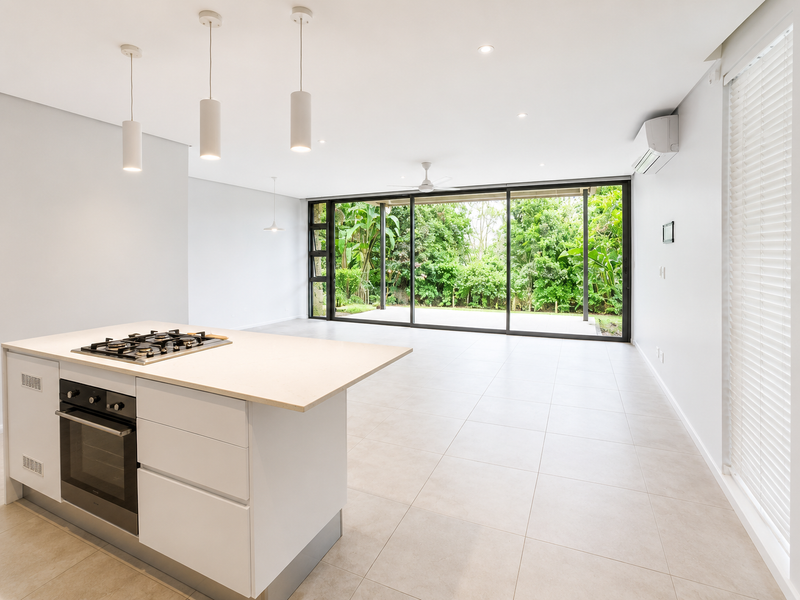 Spacious open-plan living area featuring a modern kitchen island with built-in appliances, tiled floors, and large sliding doors opening to a lush garden view.