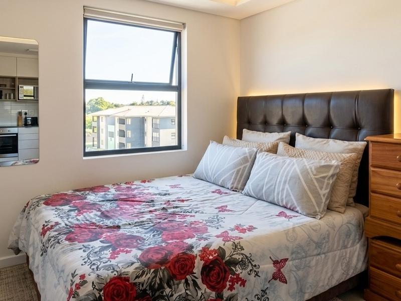 A well-maintained bedroom featuring a top-hung window with a roller blind, recessed ceiling lighting, and a wall-mounted reading light.