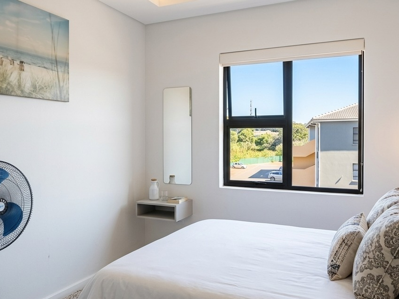 A bright, modern bedroom featuring a black aluminium window frame, roller blind, and a convenient wall-mounted floating shelf.