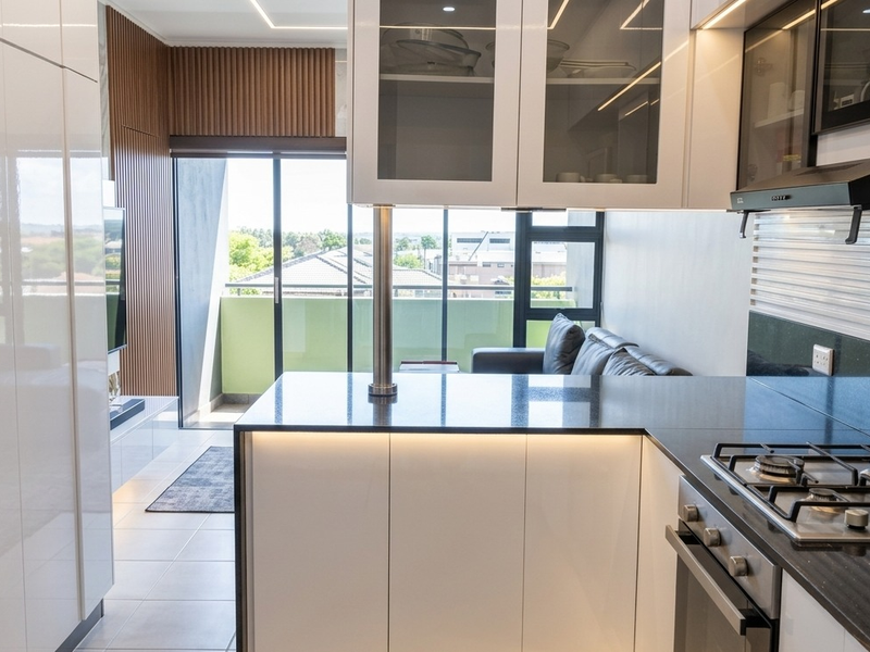 Modern open-plan kitchen featuring high-gloss cabinetry, granite countertops, and integrated gas hob, flowing seamlessly into a living area with access to a private balcony.