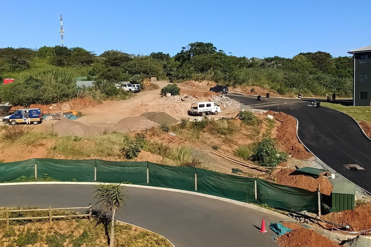 An aerial view of a residential development site showing ongoing road construction, earthworks, and proximity to an existing apartment building.
