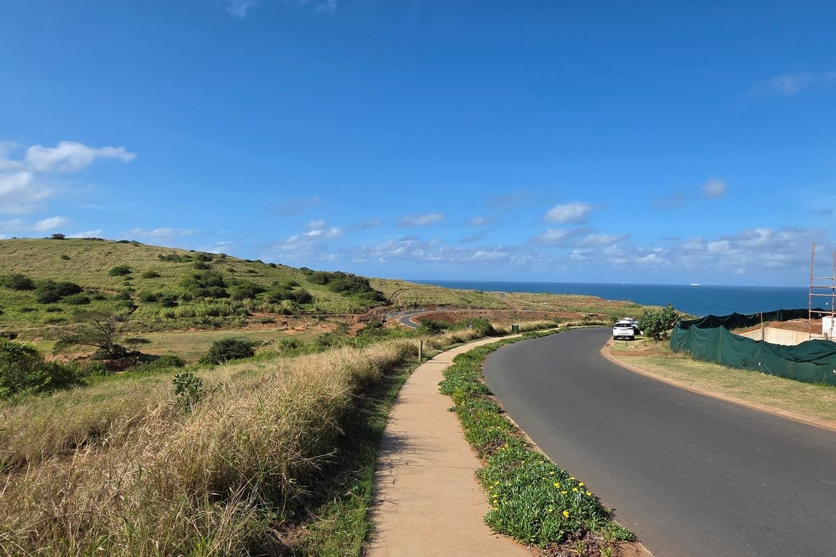 A view of a paved residential street and sidewalk leading towards a coastal landscape with ocean views.