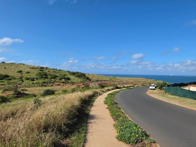 A view of a paved residential street and sidewalk leading towards a coastal landscape with ocean views.