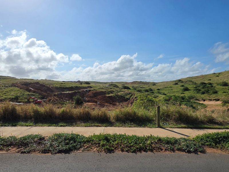 A view of a vacant residential land plot featuring a paved sidewalk and road frontage with natural hilly terrain in the background.