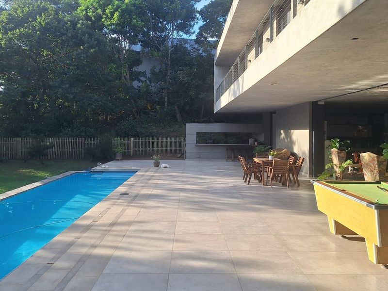 Spacious outdoor entertainment area featuring a swimming pool, large tiled patio, and a built-in braai, set against a lush garden backdrop.