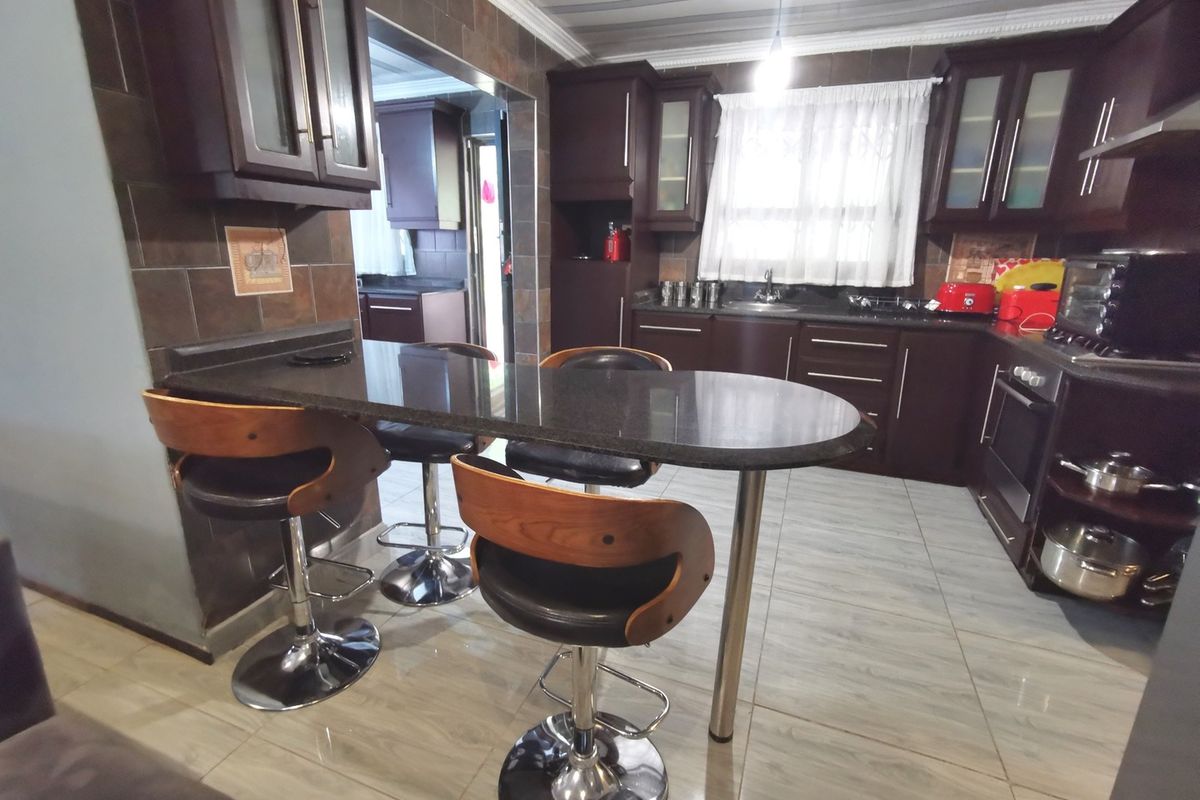A functional kitchen featuring dark wood cabinetry, granite countertops, a breakfast bar, and tiled finishes, with an adjacent scullery area visible.