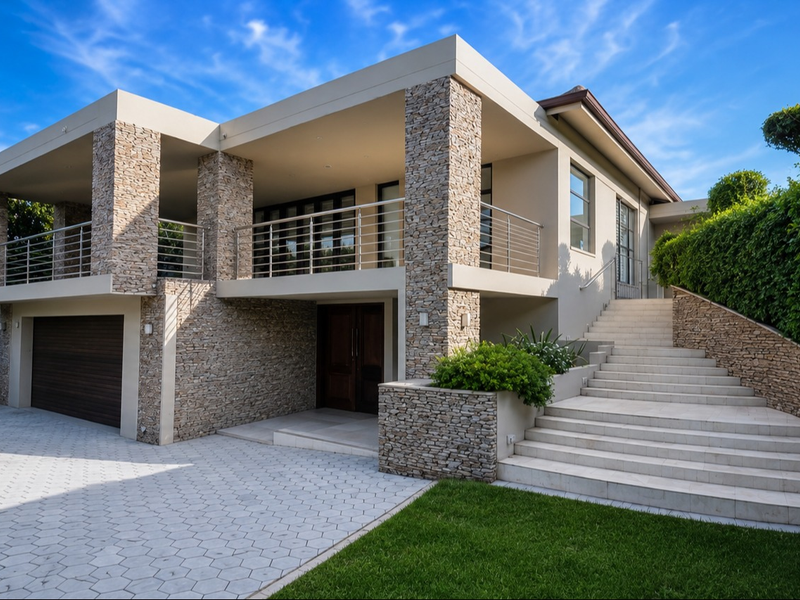 Modern multi-level residence featuring stone cladding, a spacious balcony with stainless steel balustrades, a paved driveway, and an integrated double garage.