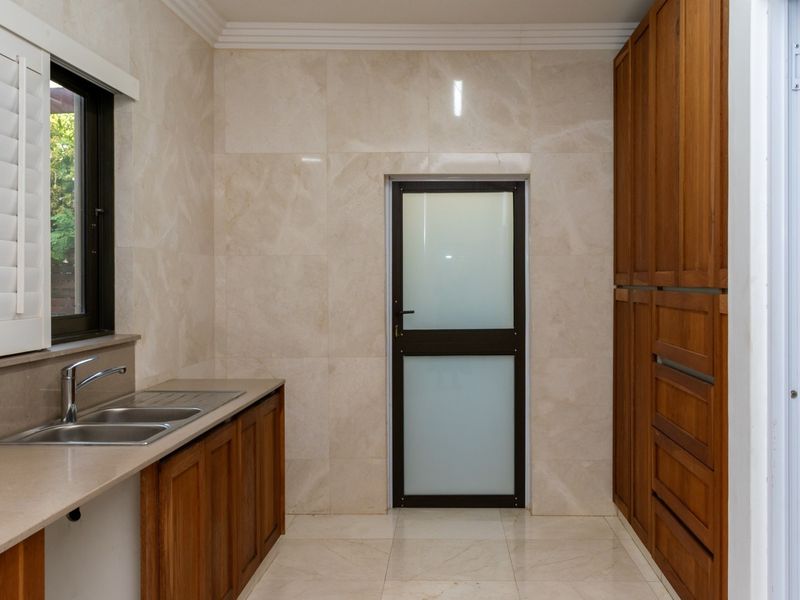A clean and functional scullery featuring built-in wooden cabinetry, stone countertops, a stainless steel double sink, and tiled finishes throughout.
