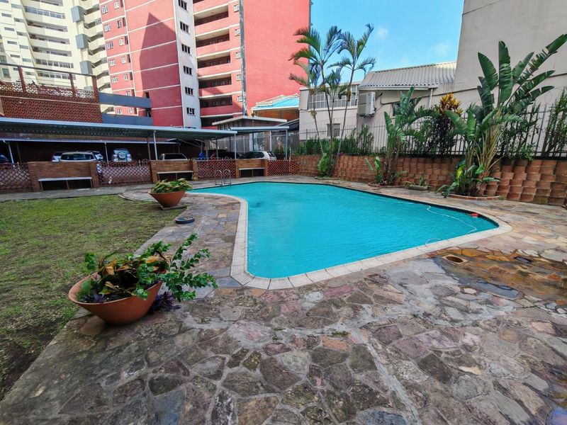 Communal swimming pool area featuring stone paving, a brick boundary wall, and adjacent covered parking structure within an apartment complex.