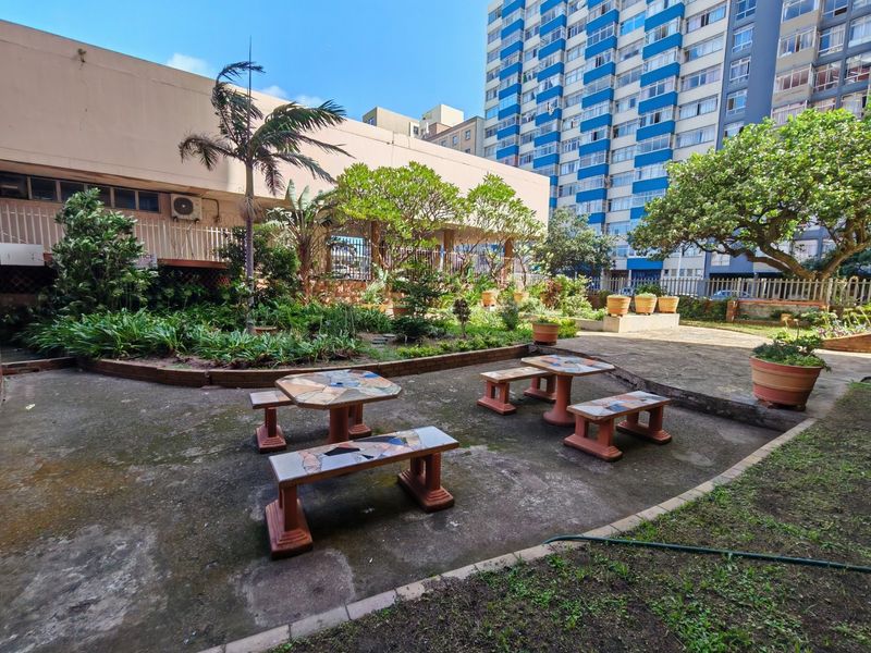A communal outdoor courtyard area featuring paved surfaces, established garden beds, and perimeter fencing within an apartment complex setting.
