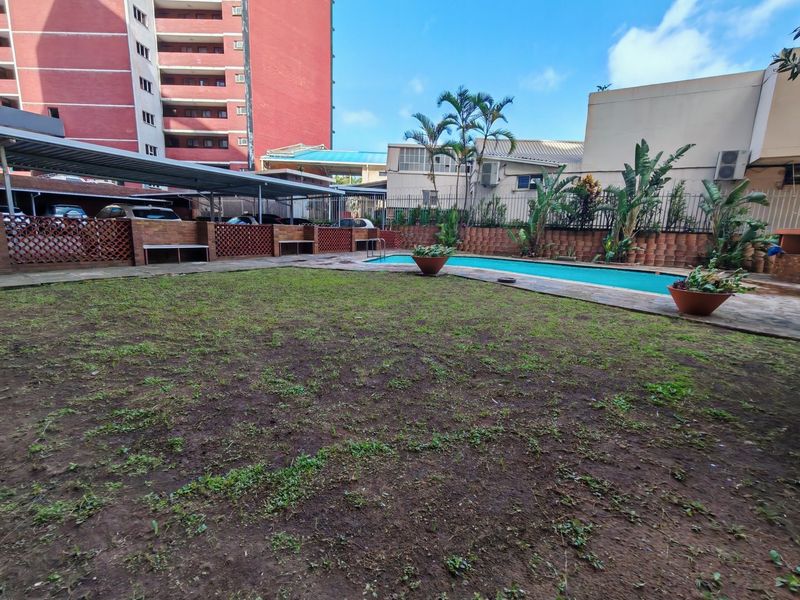 Communal outdoor area featuring a swimming pool, paved surround, and covered parking structure within a fenced complex.