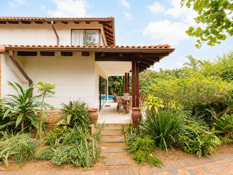 A covered outdoor patio area featuring timber beams and tiled flooring, overlooking a lush garden and swimming pool.