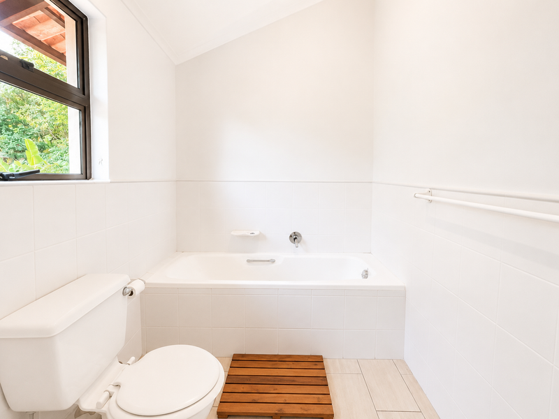 A clean, minimalist bathroom featuring a built-in bathtub, white wall tiling, and a toilet, with natural light provided by a side window.
